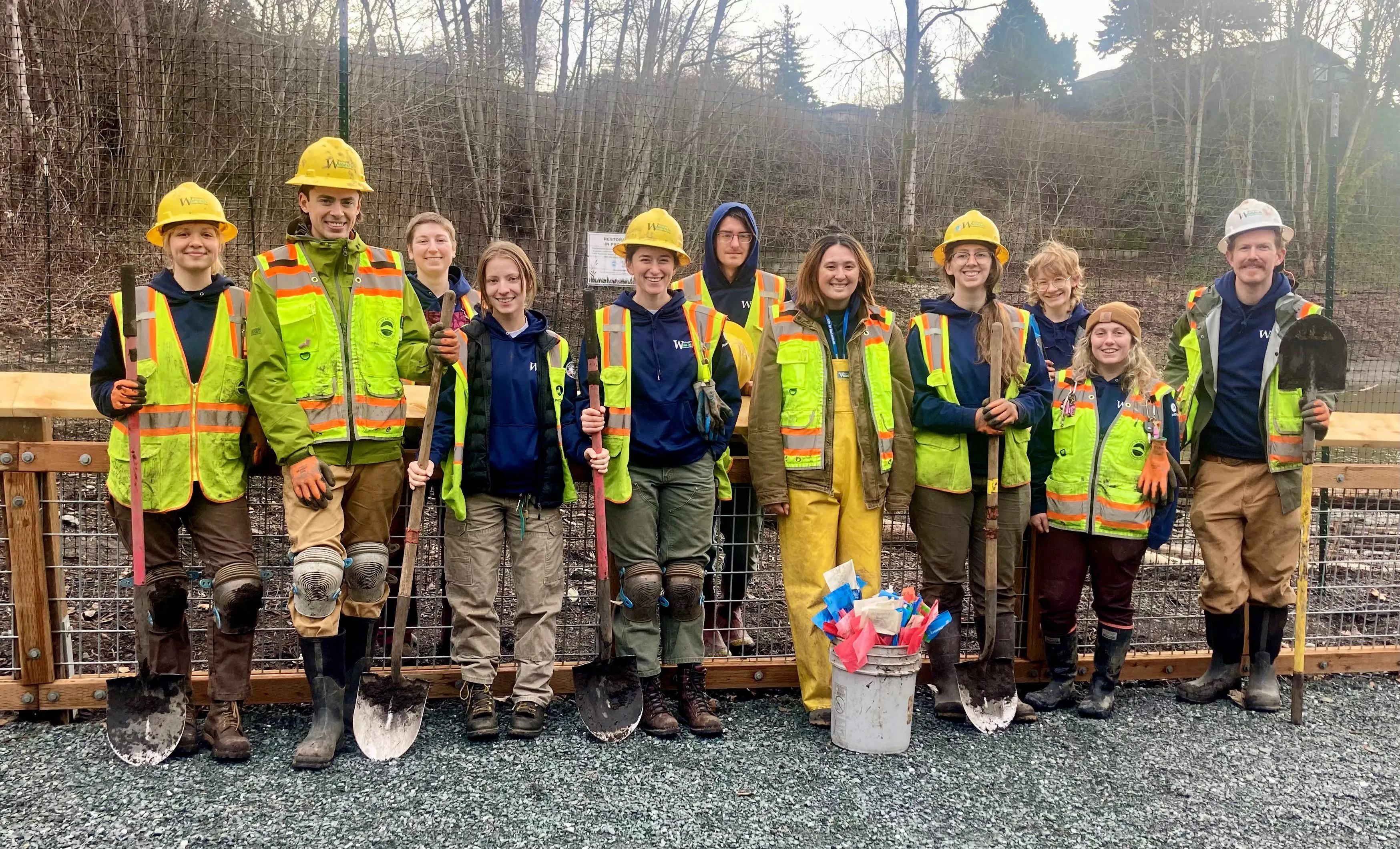 People posed in construction gear with shovels and other equipment in front of a fence