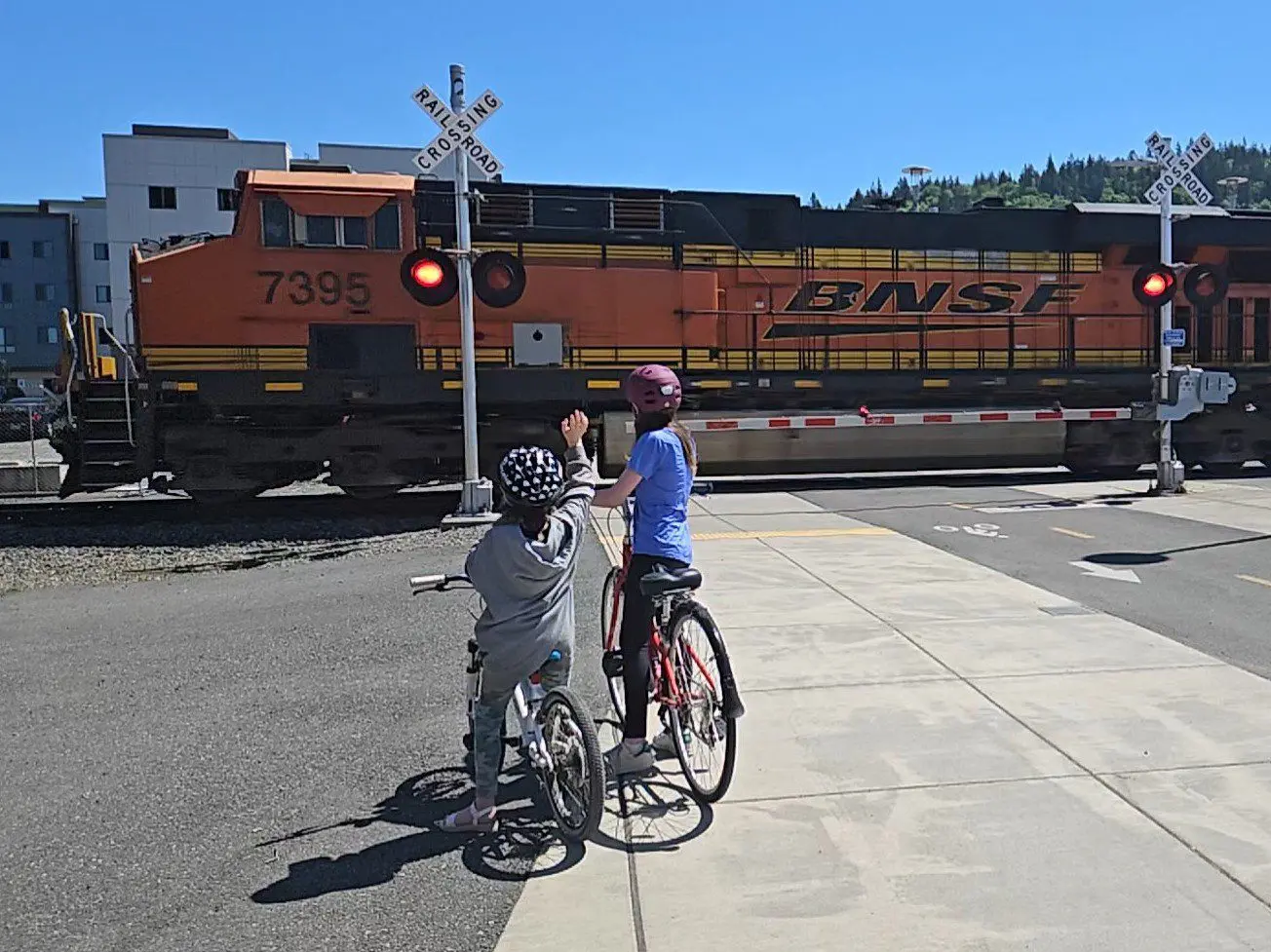 two children on bikes watching train as it travels through a street crossing