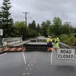 road closed sign posted in roadway near bridge with several construction workers in high visibility clothing in the background talking