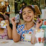 Two young girls smile while holding their craft projects.