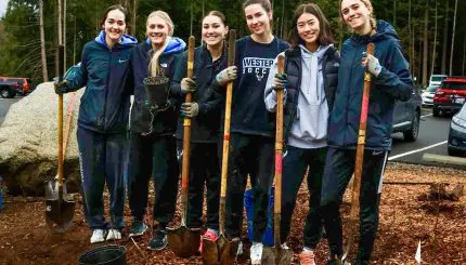 Six young adults stand shoulder to shoulder holding shovels and smiling