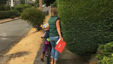 Adult walking with a child on a bike on a sidewalk trying to navigate around overgrown vegetation that is blocking sidewalk