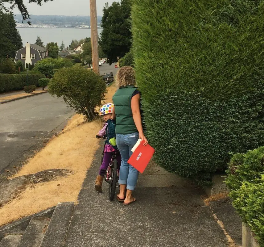 Adult walking with a child on a bike on a sidewalk trying to navigate around overgrown vegetation that is blocking sidewalk