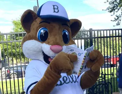 A hamster mascot with a large smile and wearing a baseball uniform reads a children's book