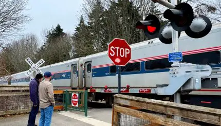 Two people stand waiting at a crossing for a train to go by