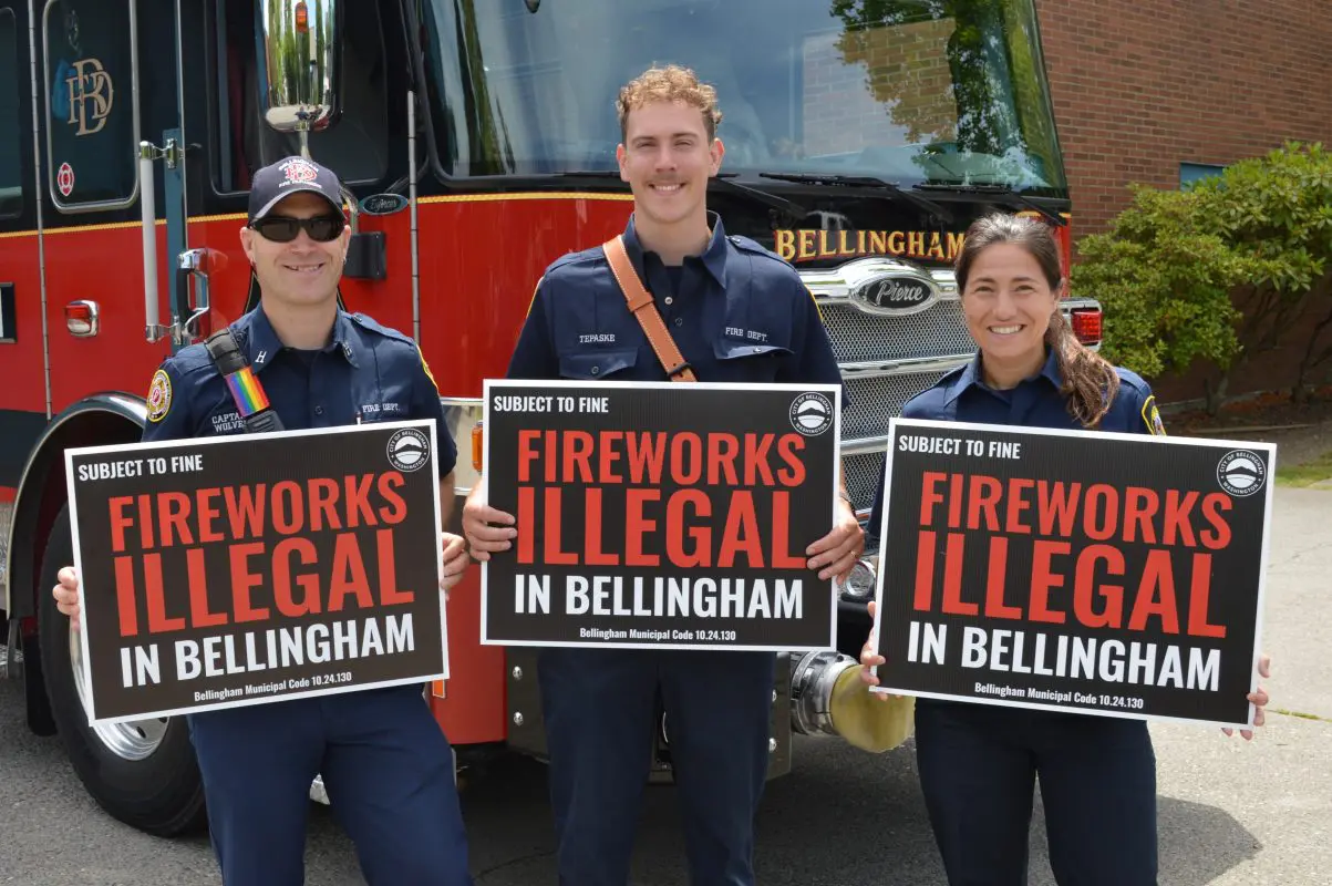 Three firefighters standing in front of a firetruck holding yard signs that read "Fireworks Illegal In Bellingham, subject to fine."