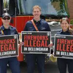 Three firefighters standing in front of a firetruck holding yard signs that read "Fireworks Illegal In Bellingham, subject to fine."