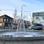 A fountain spurting water with buildings and cars in the background.