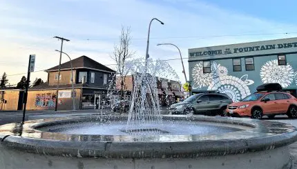 A fountain spurting water with buildings and cars in the background.