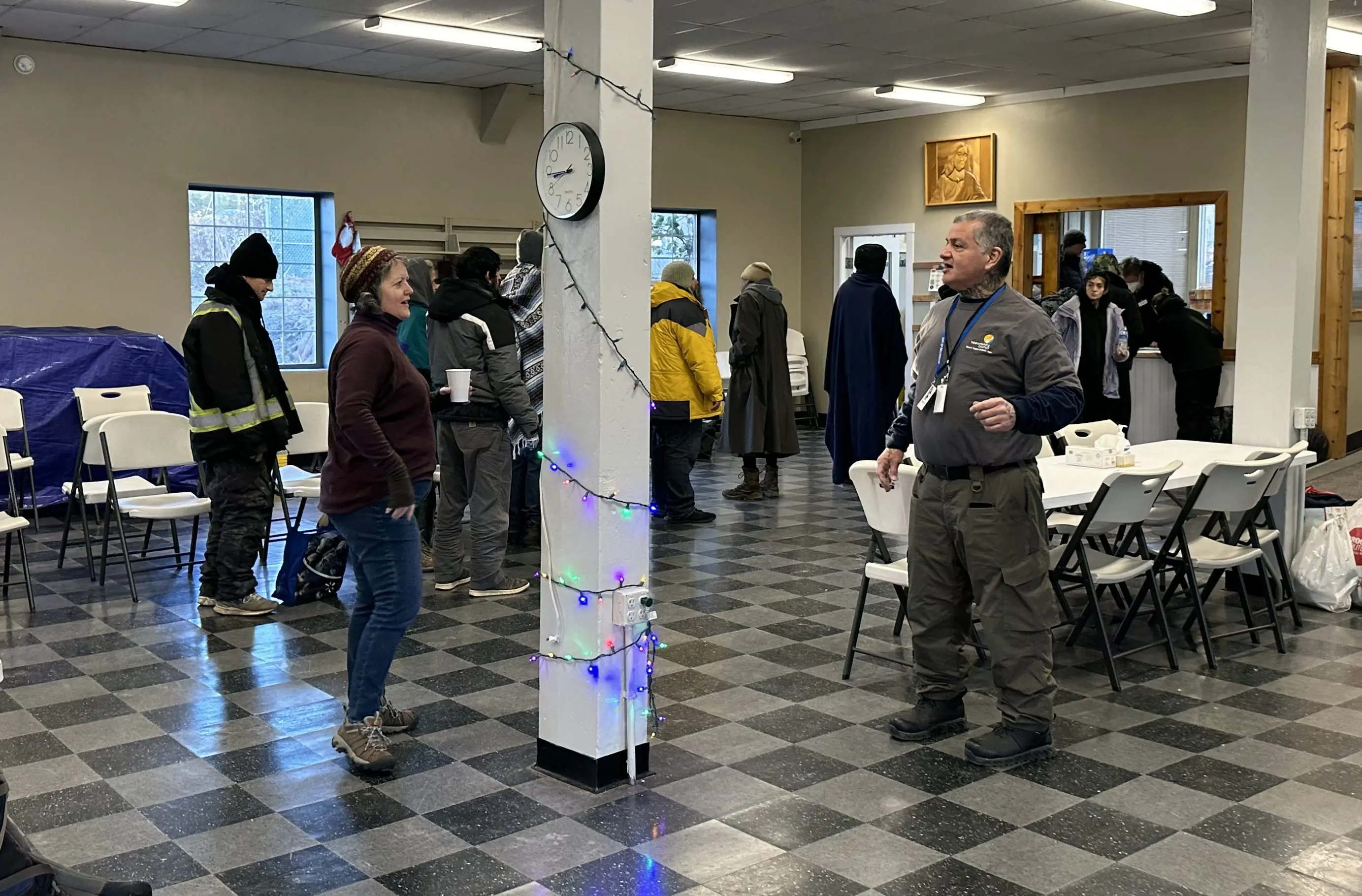 Two shelter workers talk. Shelter guests wait in a line in the background.