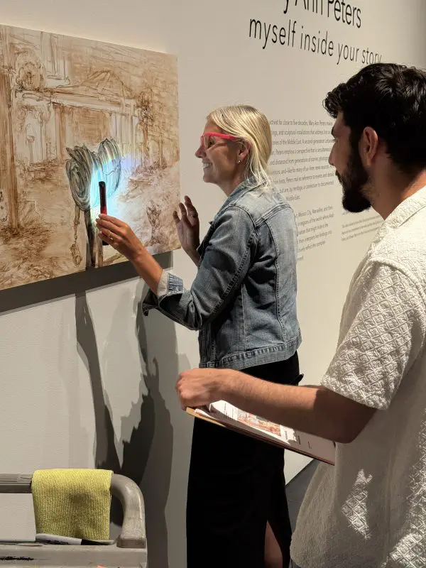 Two people examine a piece of art hanging on the wall