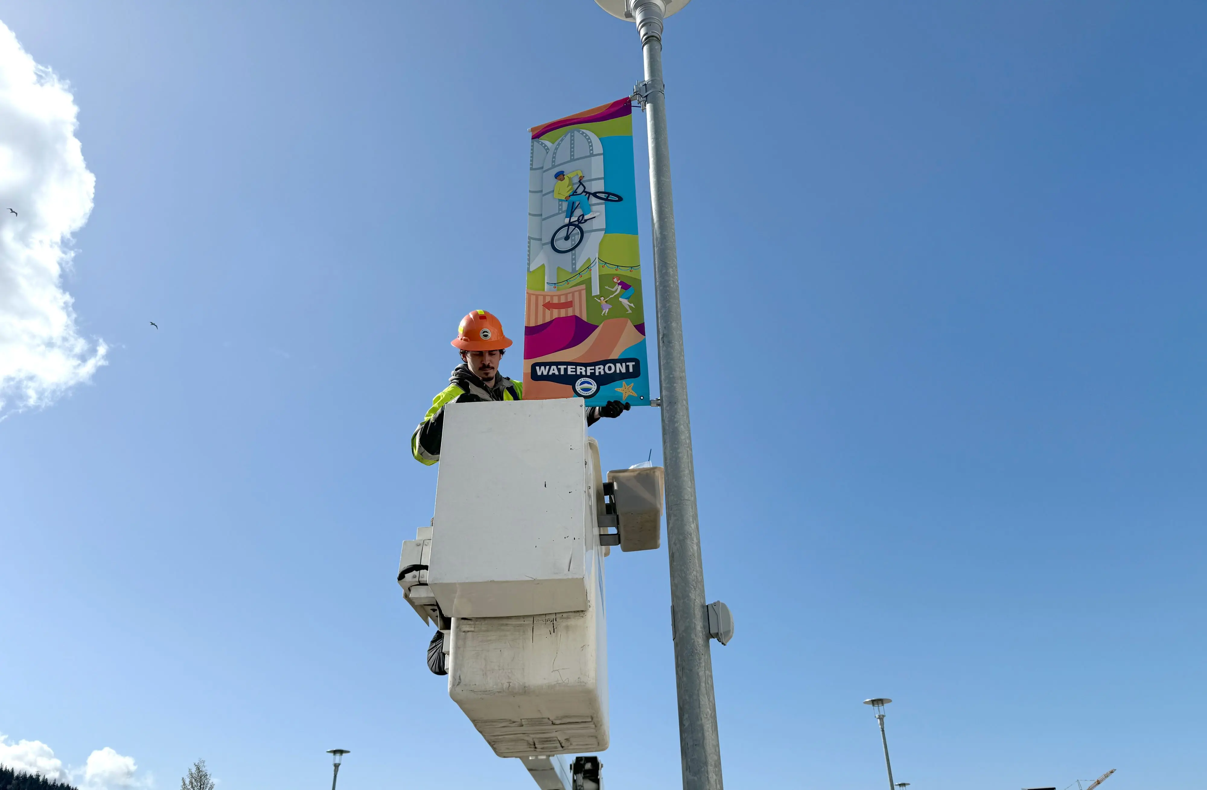 A city worker in a bucket truck installs a colorful rectangular banner in the Waterfront district.
