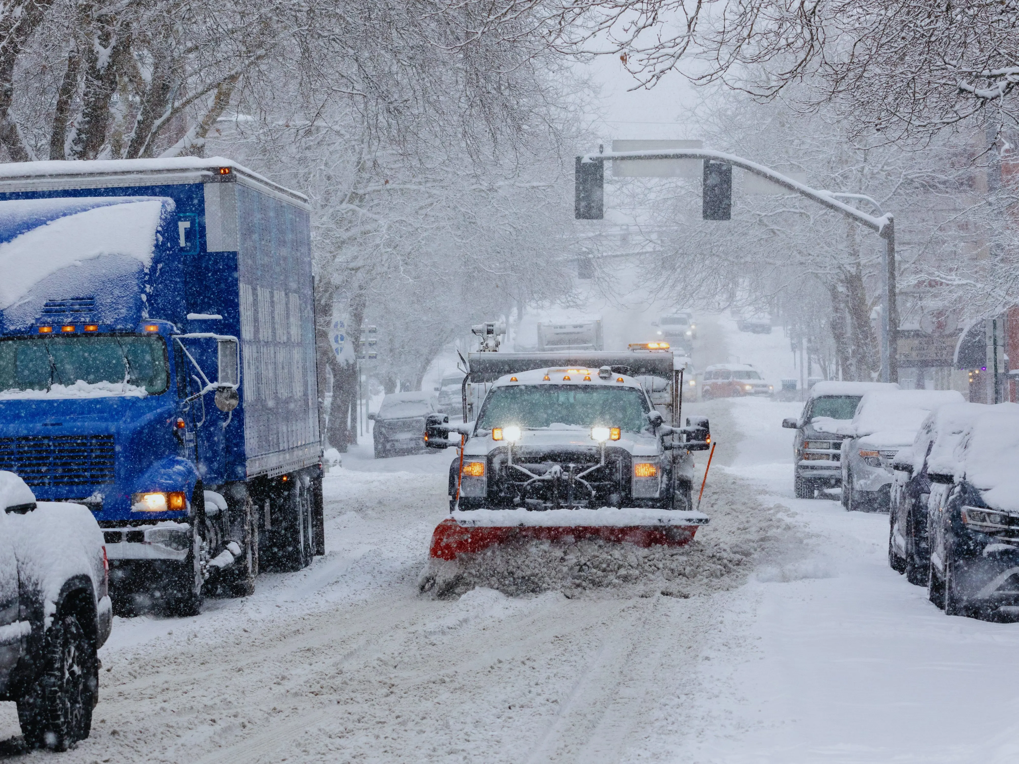 Truck with snow plow clearing snow on urban street with cars parked and driving nearby