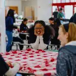 Several young people smile as they create a fleece blanket together around a round table.