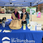 Two women smile with a basket of tree saplings in paper bags on a table