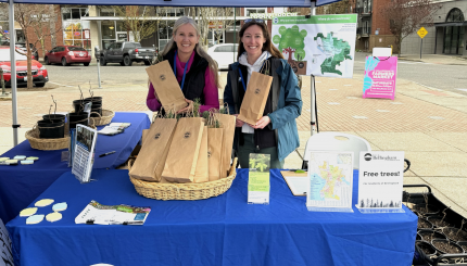 Two women smile with a basket of tree saplings in paper bags on a table