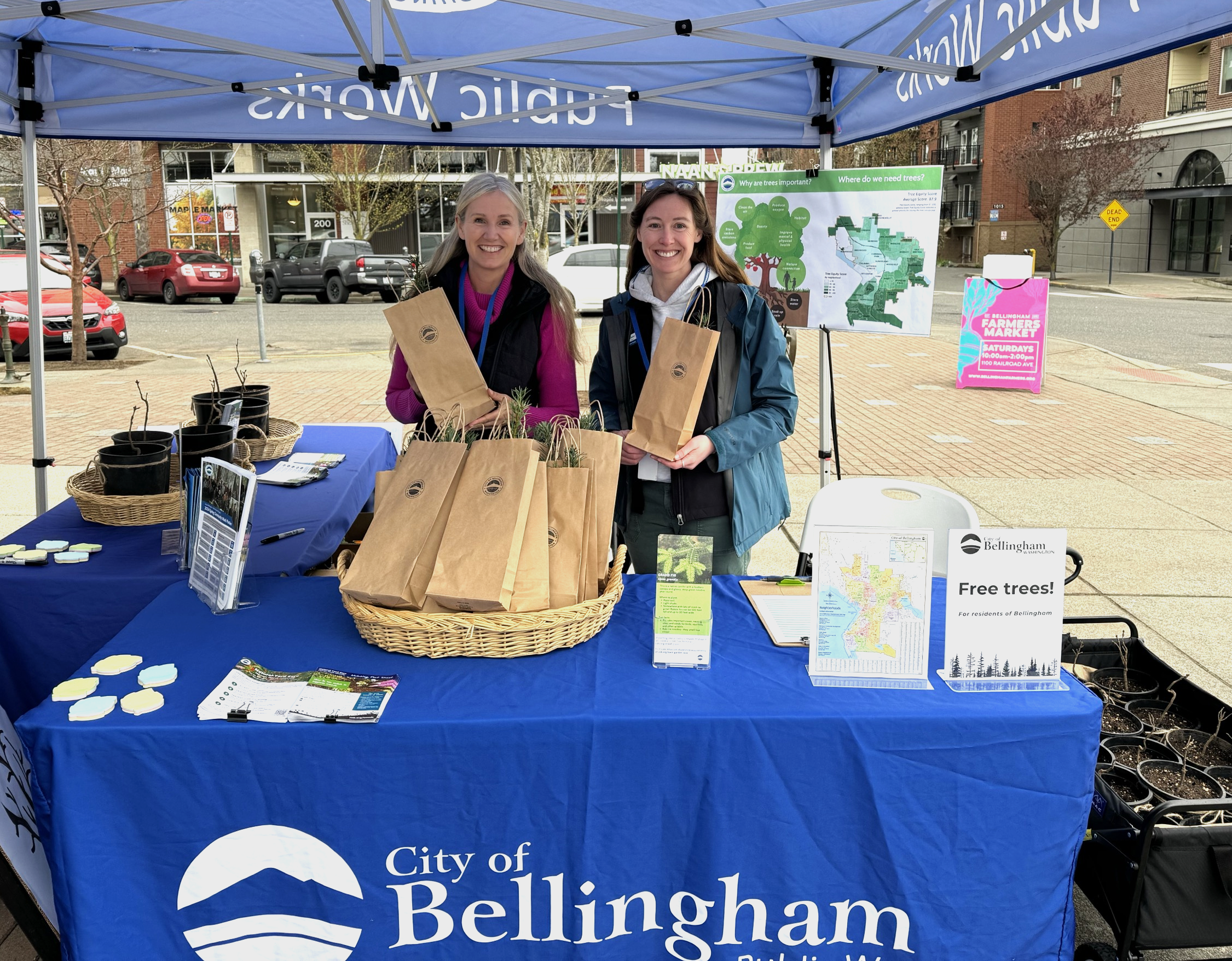 Two women smile with a basket of tree saplings in paper bags on a table