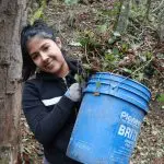 Young adult smiling, standing in forest, holding blue bucket overflowing with weeds
