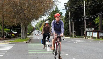 Two bike riders with helmets riding in a bike lane on a two-lane road through suburban area.