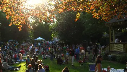 A large crowd gathers on a sunny park lawn to listen to music.