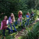 A row of five kids smile as they help remove ivy from a park.