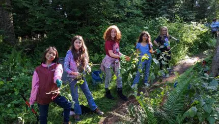 A row of five kids smile as they help remove ivy from a park.