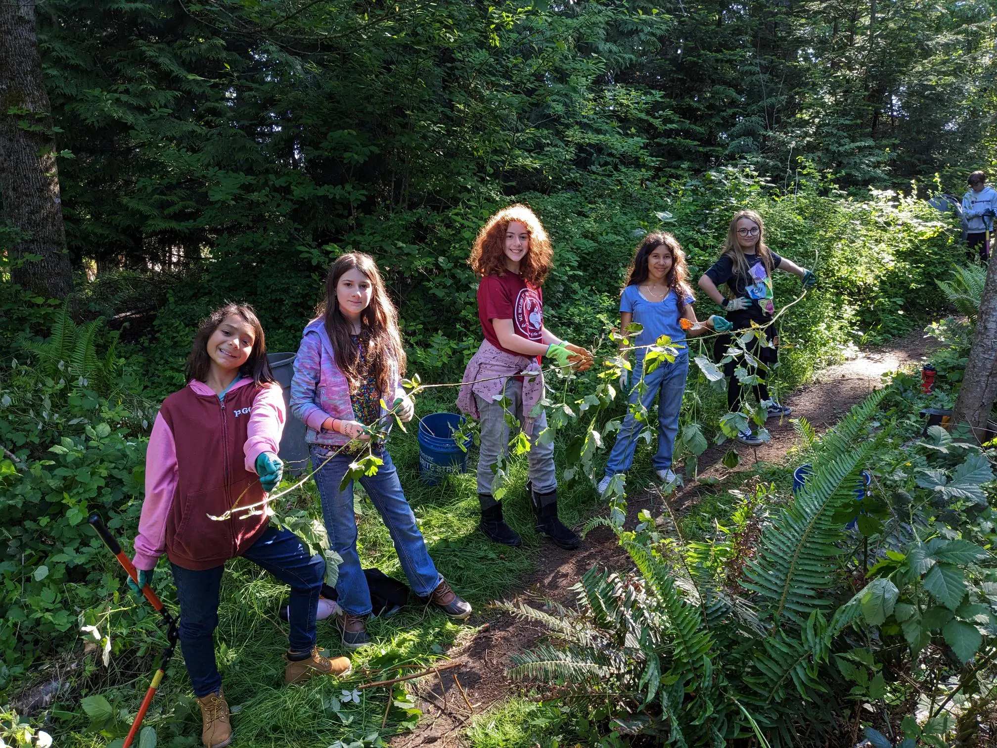 A row of five kids smile as they help remove ivy from a park.