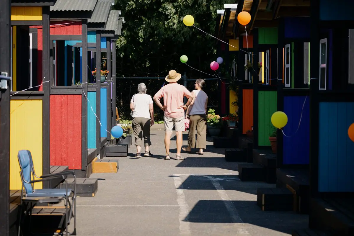 Three people walk through a row of colorful tiny homes
