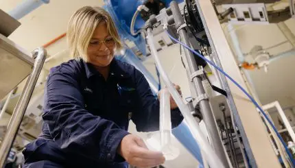 A blonde woman in glasses and a dark blue work jumpsuit, smiles while carefully working with clear tubes and equipment in what appears to be a water treatment facility. Large blue pipes are visible in the background.