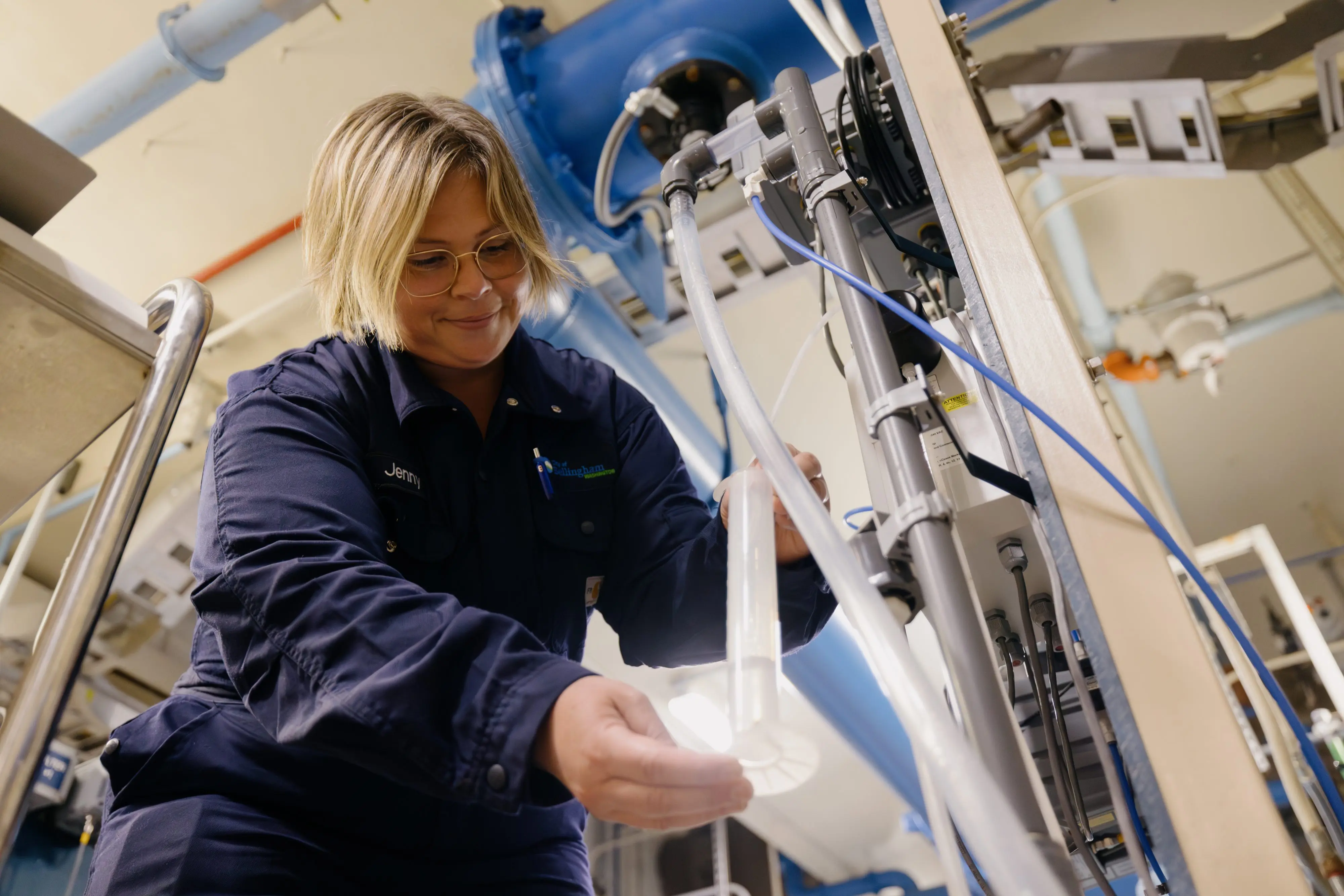 A blonde woman in glasses and a dark blue work jumpsuit, smiles while carefully working with clear tubes and equipment in what appears to be a water treatment facility. Large blue pipes are visible in the background.