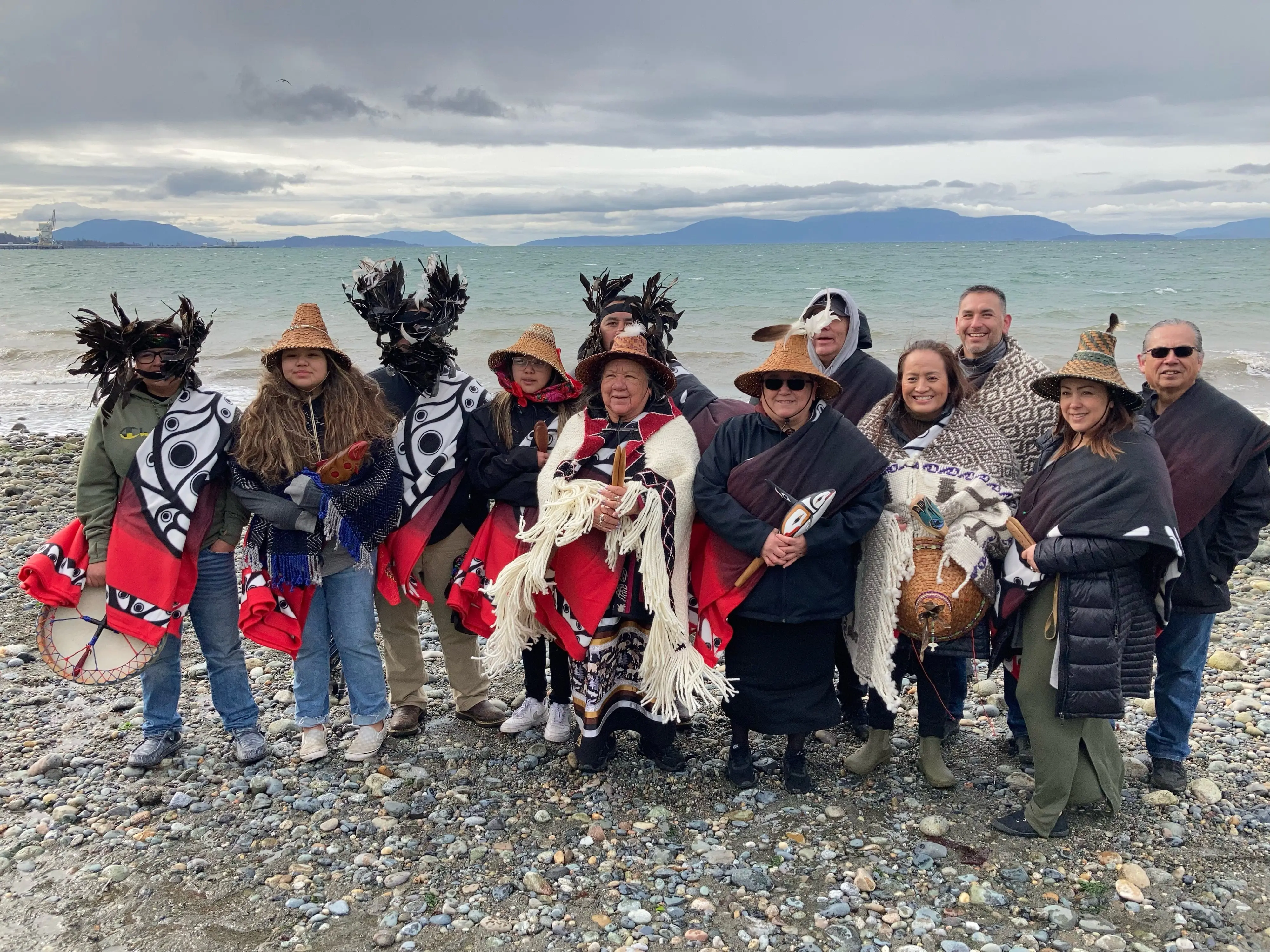 Group of individuals in traditional Native American attire standing on cobbled beach