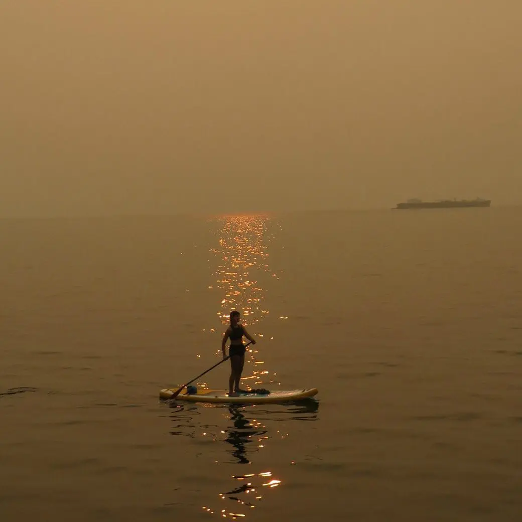 Person standing on a paddleboard in water with sun shining through a smoky sky.