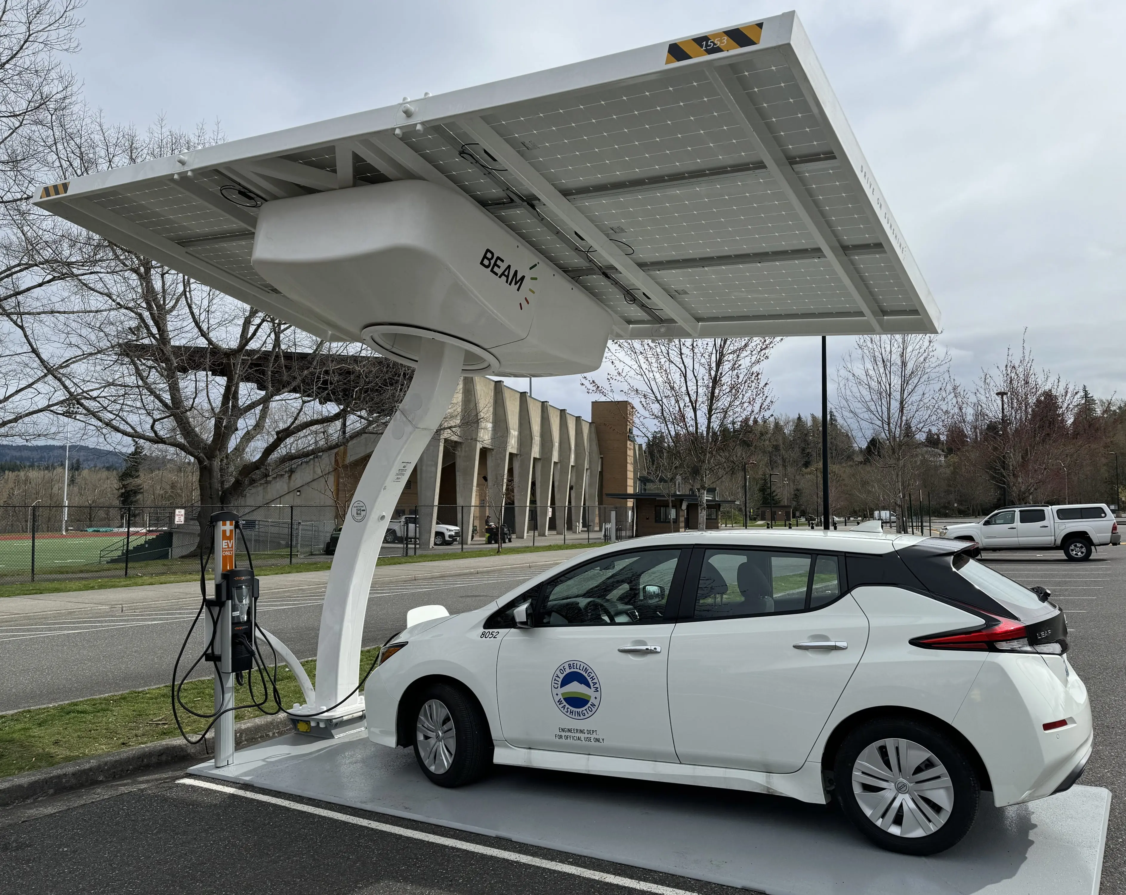 Small passanger vehicle plugged into charger with large solar panel over head.