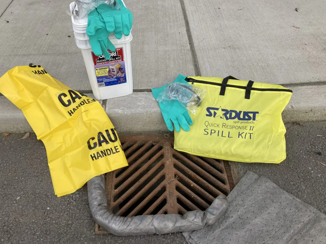 Quick response spill kit with a bucket, gloves, goggles, and other materials next to an outdoor storm drain