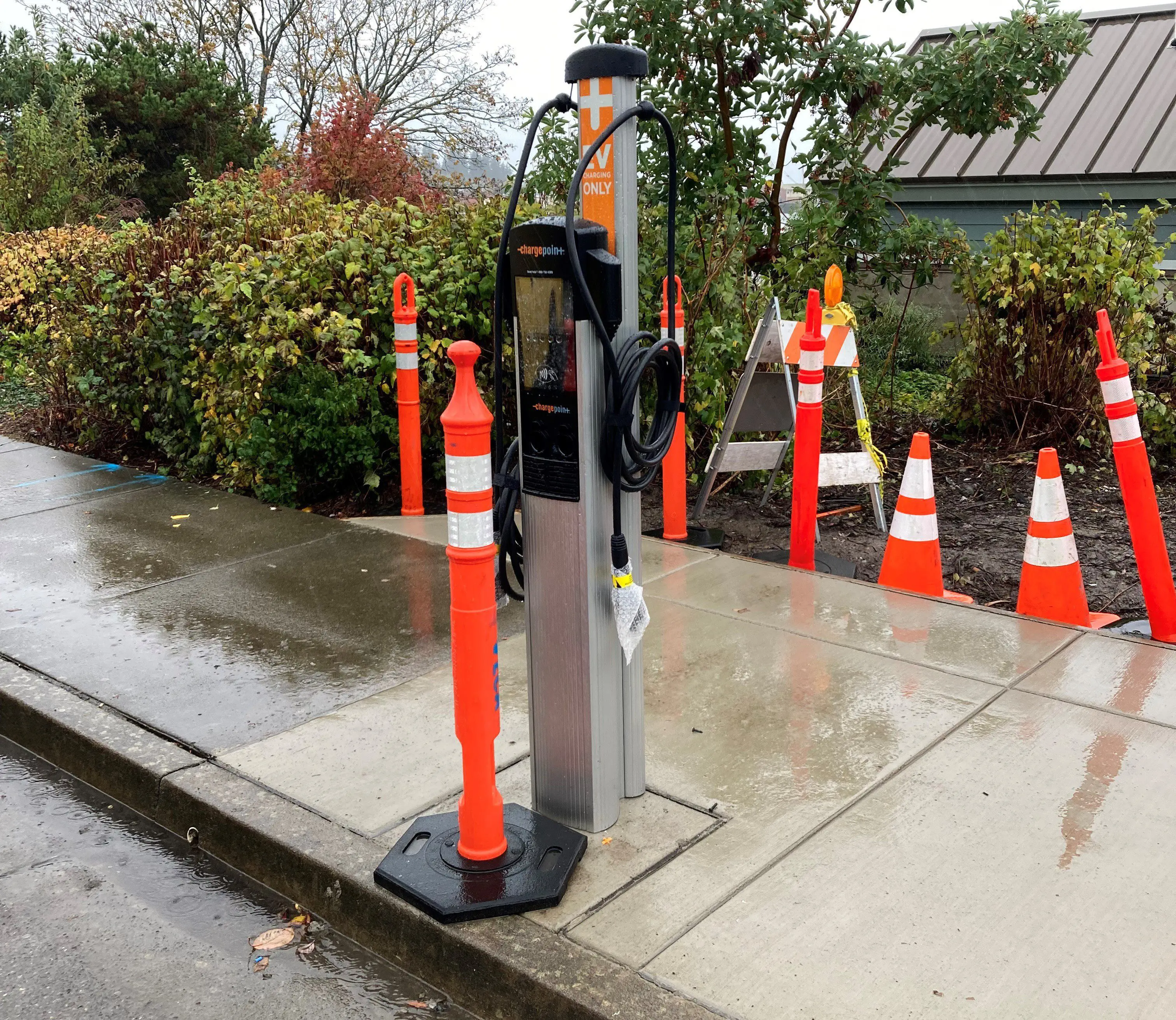 New electric vehicle charging station along road on a rainy day. Orange cone in front of station to block use.