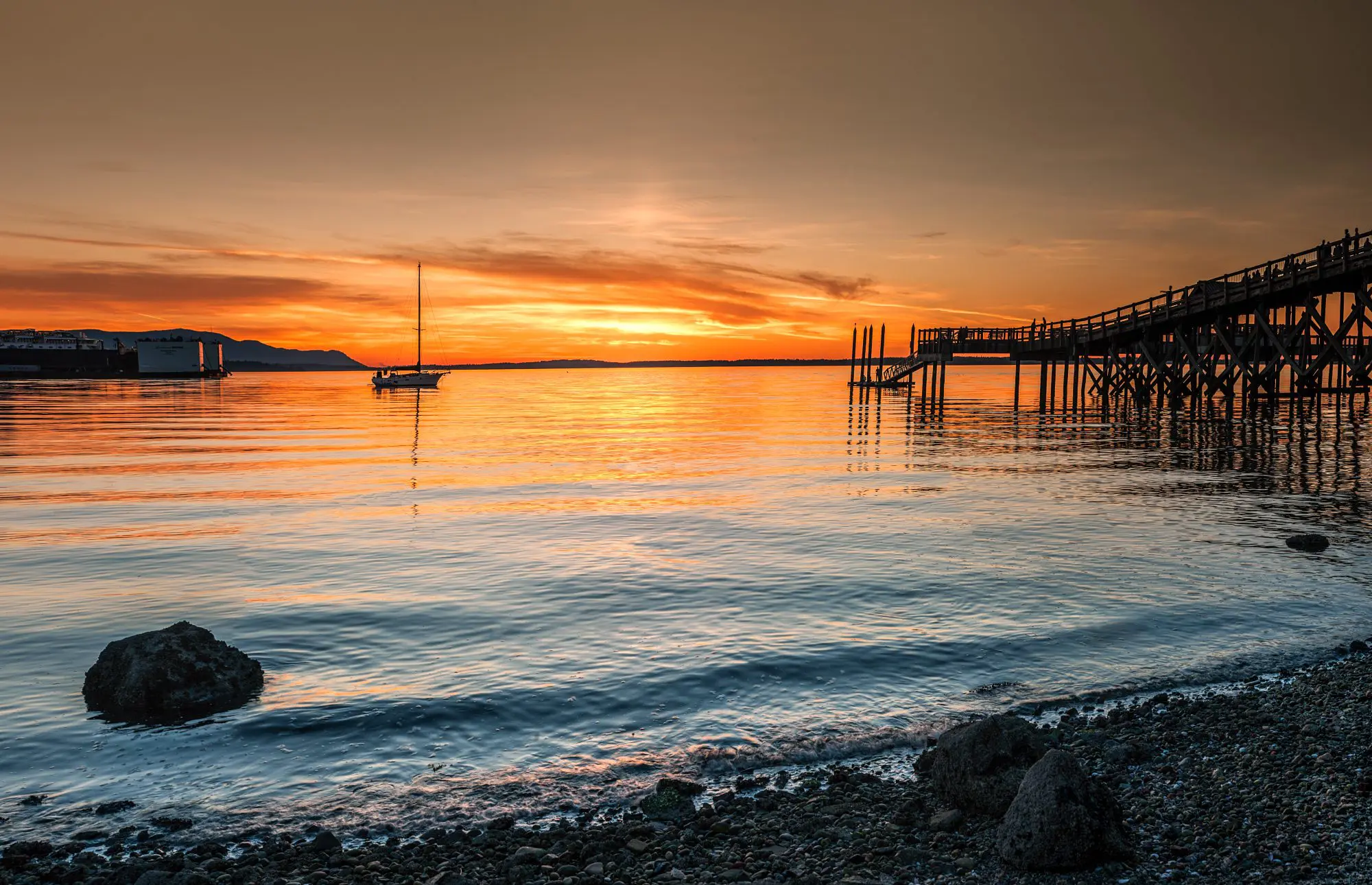 Sunsetting over marine waters with sailboat anchored in distance and large dock on the side