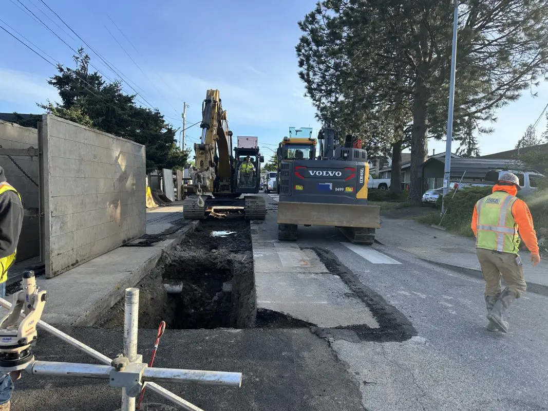 Trench in middle of road with construction worker operating equipment nearby.