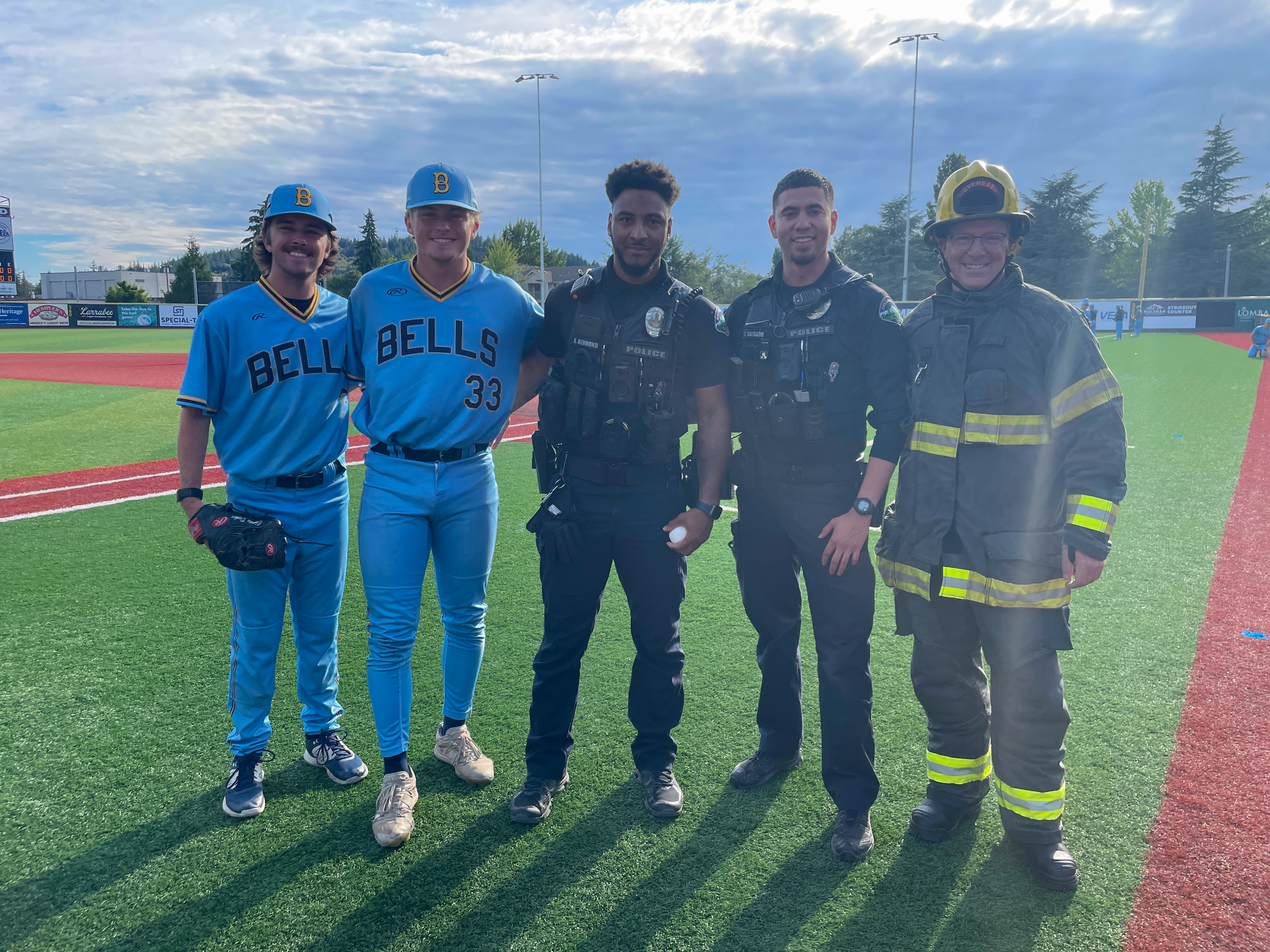 Two baseball players, two police officers, and a fire fighter smile together on a baseball field