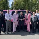 Seven police officers, two in pink suites, pose in front of a patrol car wrapped in a pink and purple design.