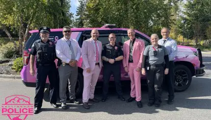 Seven police officers, two in pink suites, pose in front of a patrol car wrapped in a pink and purple design.