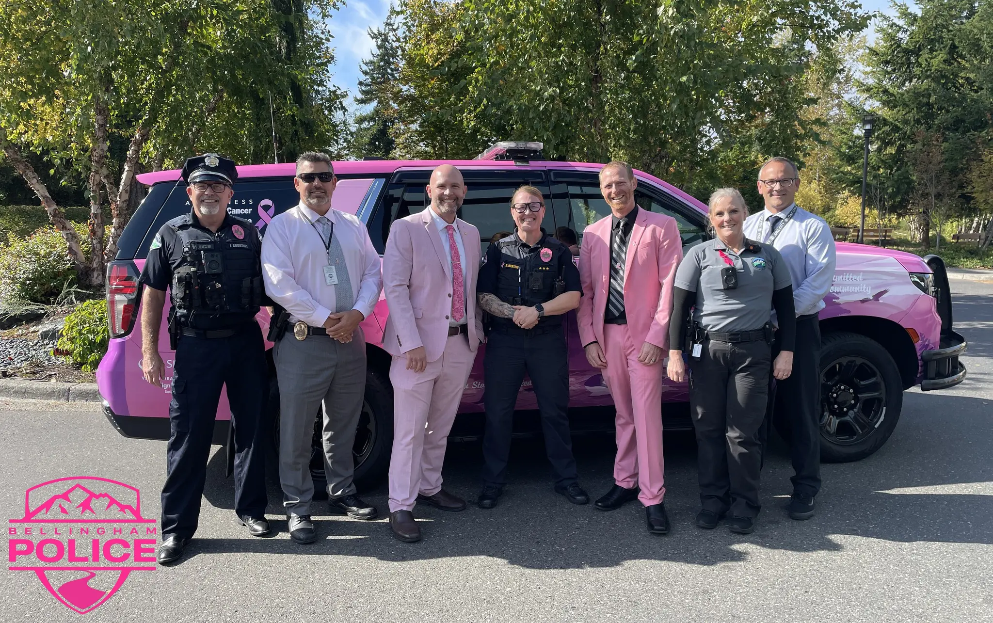 Seven police officers, two in pink suites, pose in front of a patrol car wrapped in a pink and purple design.