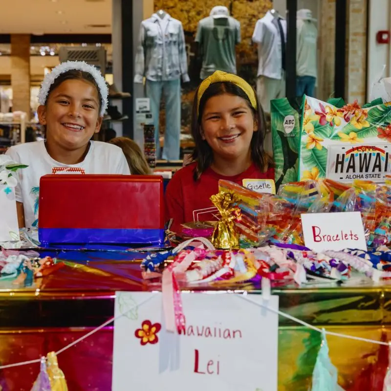 Two children smile behind a table selling leis