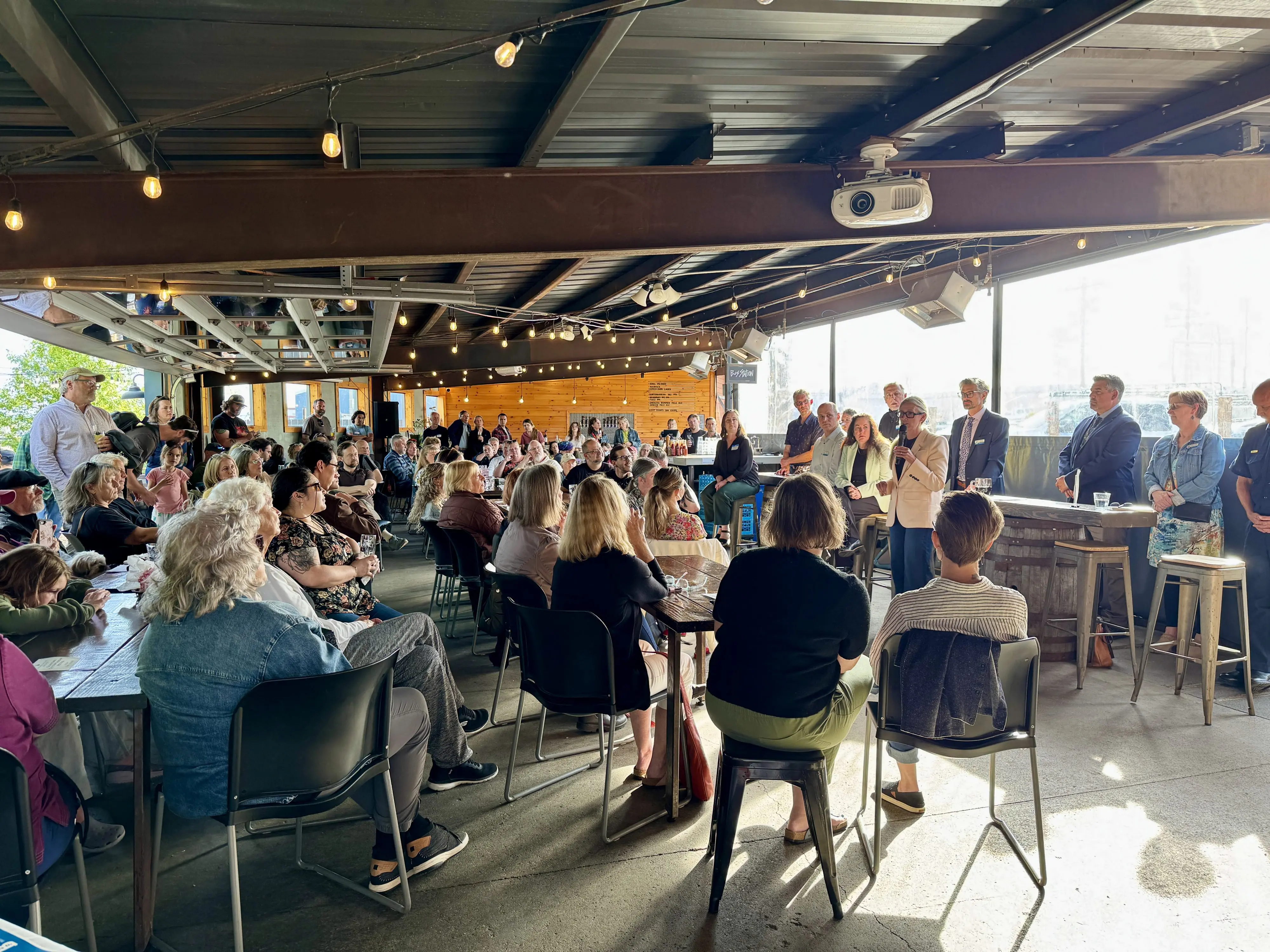 City leaders speak to a large crowd in a brewery.