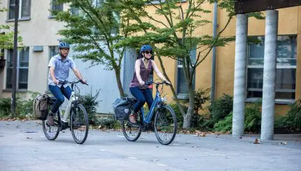 Two people riding ebikes in front of a building. 