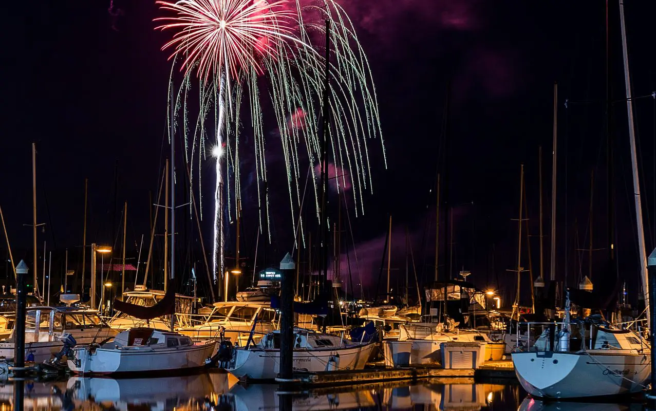 Fireworks explode over boats in a harbor.