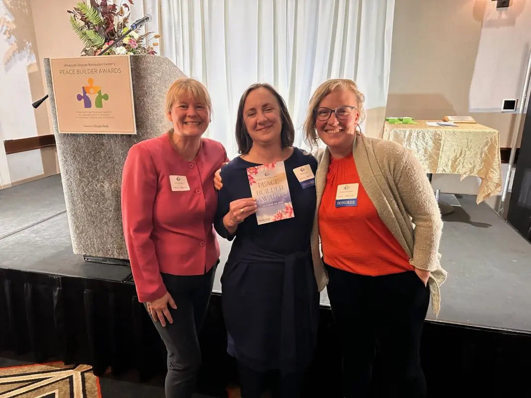 Three women pose with an award in front of a stage