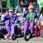 Four women dressed in festive purple and green watch a parade.