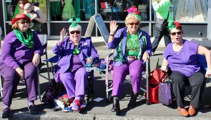 Four women dressed in festive purple and green watch a parade.