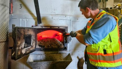 Person in yellow and orange safety vest with long pole extended into an opening in a large hearth with firey orange heat and ash