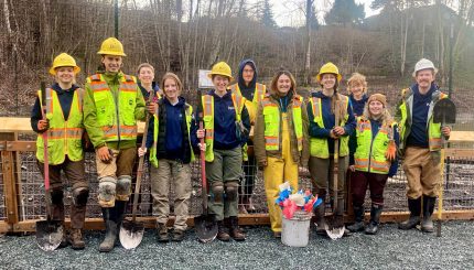 People posed in construction gear with shovels and other equipment in front of a fence